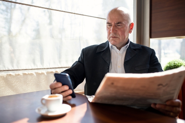 An older, bald white man wearing a smart suit, looks angrily at the mobile phone he is holding in his right hand. In his left hand is a newspaper. An espresso is on the table in front of where he is sat.