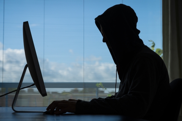 Silhouette of a person in a hoody in front of a computer screen. It is photographed from the side, against landscape and sky outside a window.