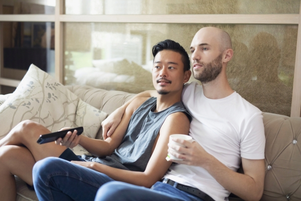 A couple sit cuddling on the sofa while watching television. The man on the left is Asian and wears a vest and short. He is holding a remote control. The man on the right is white with a shaved head and dark beard. He wears a t-shirt and jeans.