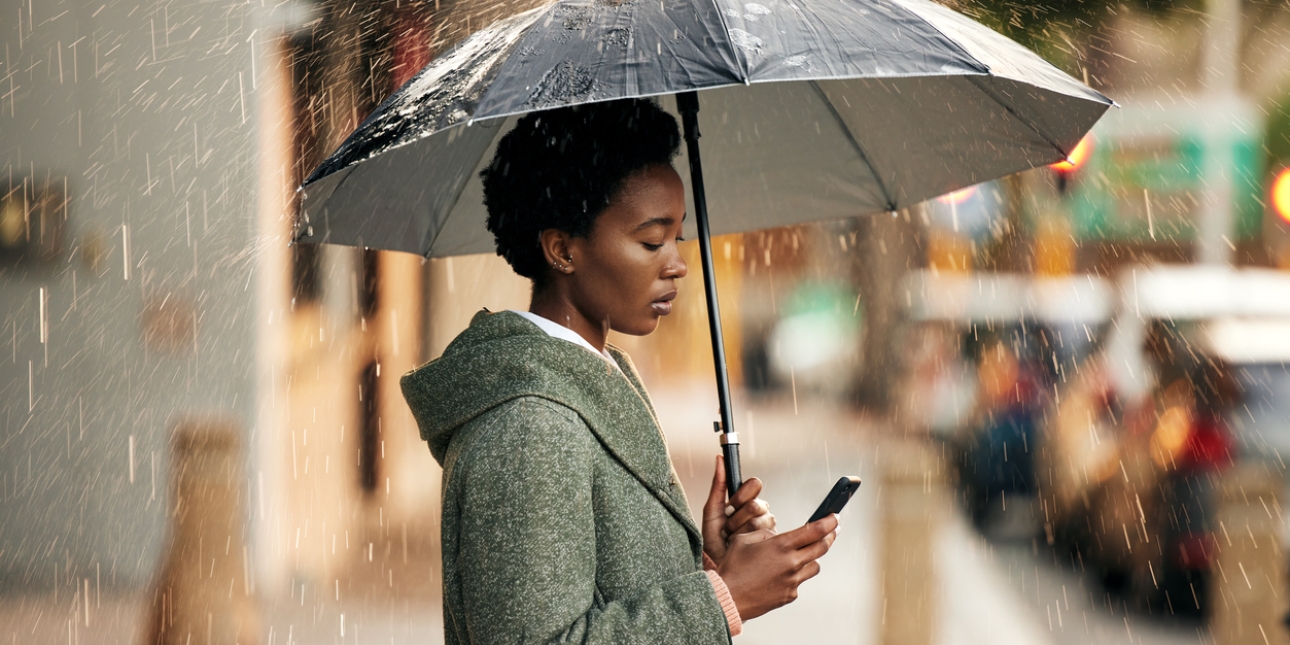 Side profile of a Black woman in a grey coat stood under an umbrella in the rain while looking at the mobile phone in her right hand