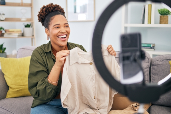 A black woman, who is sat on a sofa, laughs while holding a jacket in front of a mobile phone and ring light. In the background are a cushion and plants, books and a clock on a wooden shelving unit.