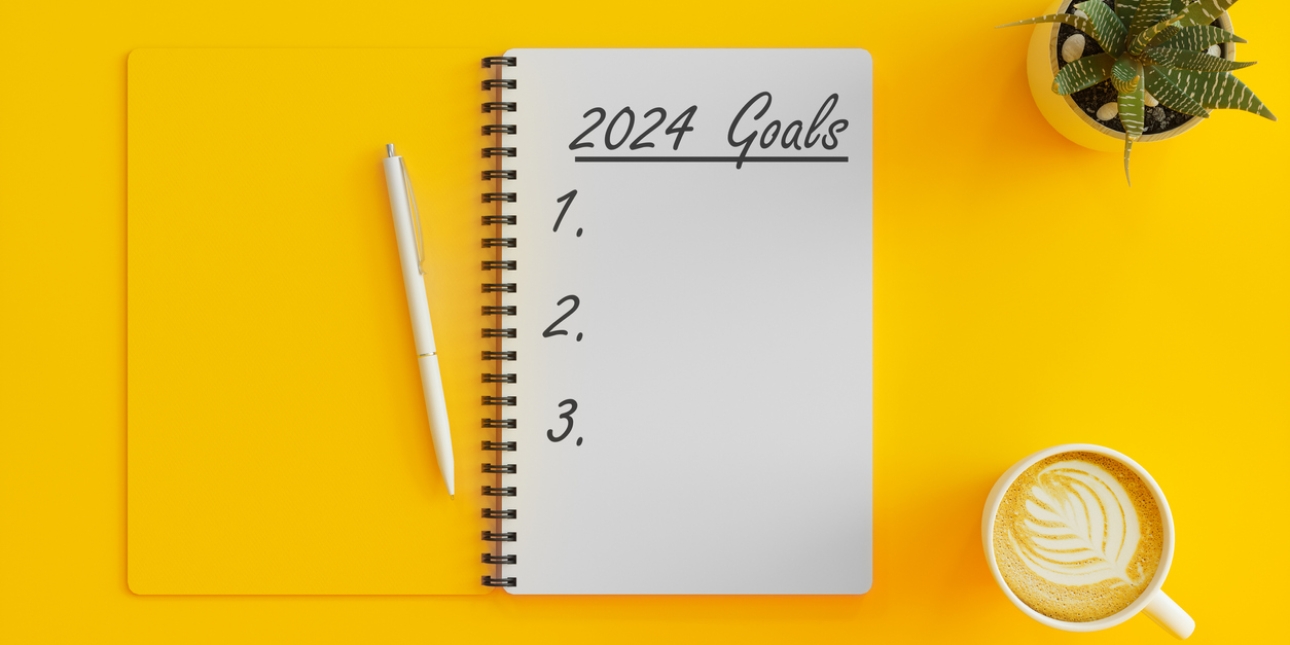 A bird's eye view of an open notebook with yellow cover and white first page, on which in handwritten 2024 goals 1, 2, 3. A pen is placed on the notebook. Next to it, on a yellow background are a cup of coffee and plant