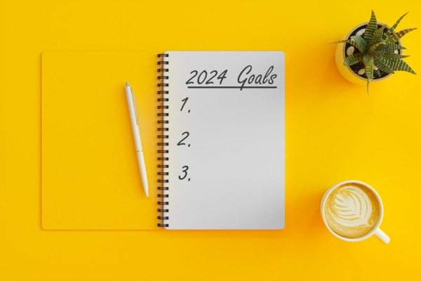 A bird's eye view of an open notebook with yellow cover and white first page, on which in handwritten 2024 goals 1, 2, 3. A pen is placed on the notebook. Next to it, on a yellow background are a cup of coffee and plant