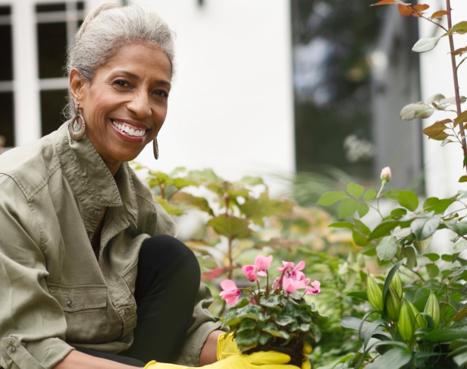 A smiling, older Black woman with grey hair, hooped earrings and khaki jacket, kneels down in her garden. Wearing yellow gloves, she is planting a pink flowering plant. Around her are green bushes a wooden bench and a house