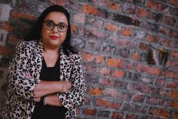 Advita Patel, a British Asian woman with dark shoulder-length hair, glasses, red lipstick, stood in front of a brick wall with her arms crossed. She wears a leopard print jacket over a black blouse.